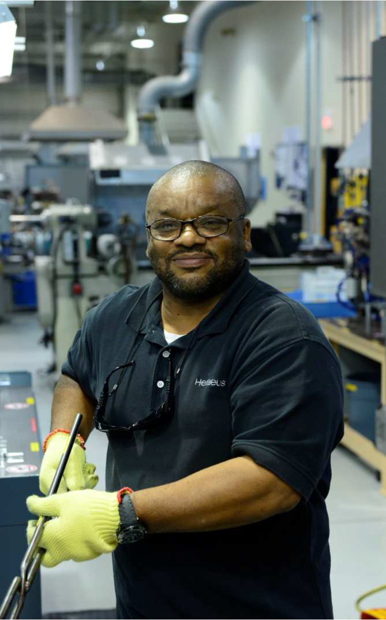 A man in work clothes and protective gloves is standing in a production hall and looking at the camera.