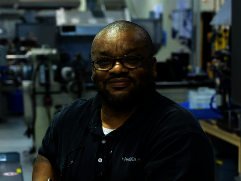 A man in work clothes and protective gloves is standing in a production hall and looking at the camera.